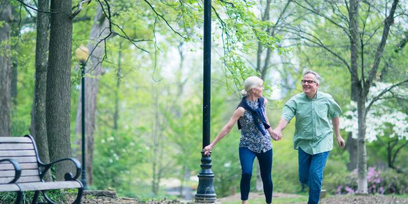An older couple holding hands and walking happily in a park surrounded by green trees.